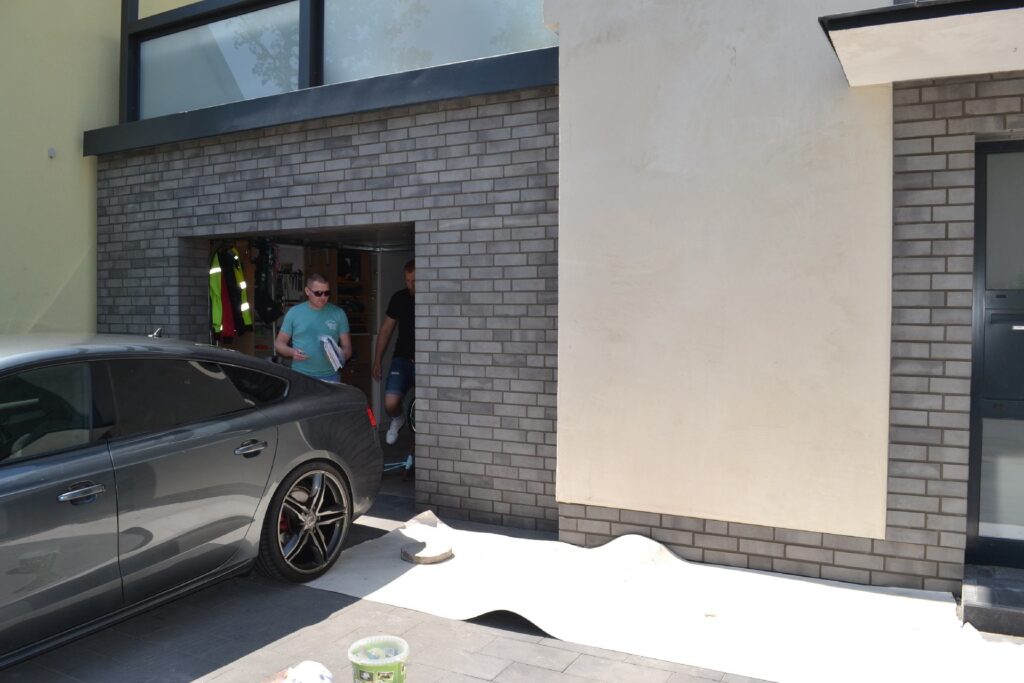 Ein Mann steht vor einer modernen Hausfassade mit Klinkerriemchen-Verkleidung neben einem grauen Auto in der Einfahrt.