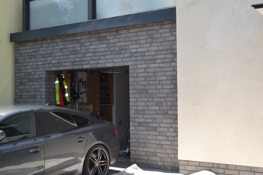 Modernes Haus mit grauer Klinkerriemchenfassade und offener Garage, in der ein dunkler PKW steht.