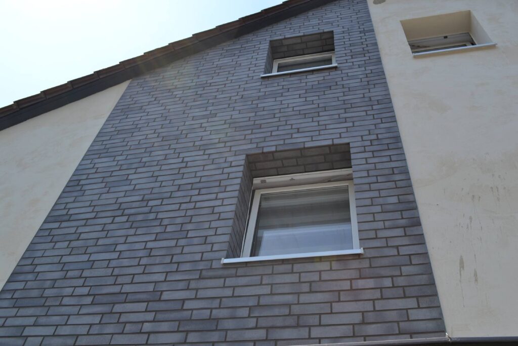 Moderne Hausfassade mit grauer Klinkerriemchenfassade und weißen Fenstern an einem zweistöckigen Wohnhaus.