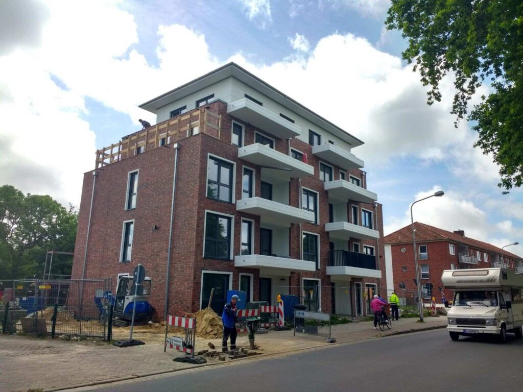 Neubau mit Klinkerriemchenfassade und weißen Balkonen, Bauarbeiten an der Straße, bewölkter Himmel im Hintergrund