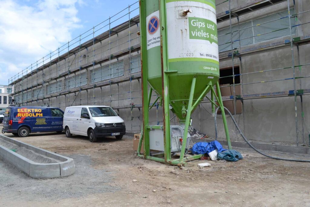 Baustelle mit Gerüst an Hausfassade, grüner Gipssilo und Fahrzeuge auf unbefestigtem Untergrund
