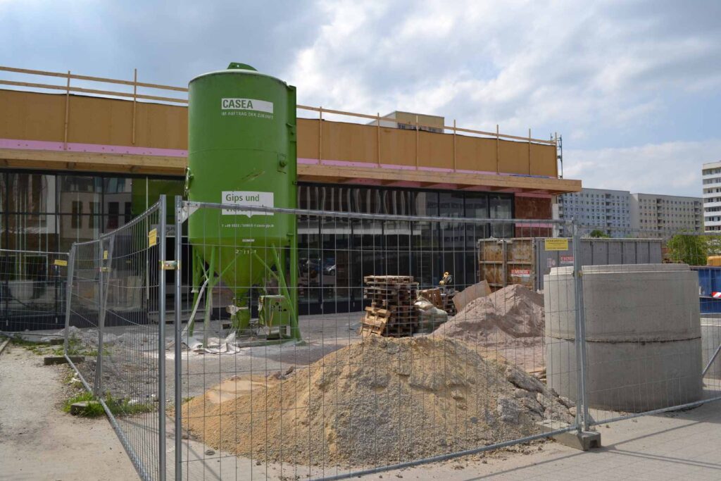 Baustelle mit grünem Silo und Rohbau eines Gebäudes mit unverkleideter Fassade, umgeben von Bauzaun