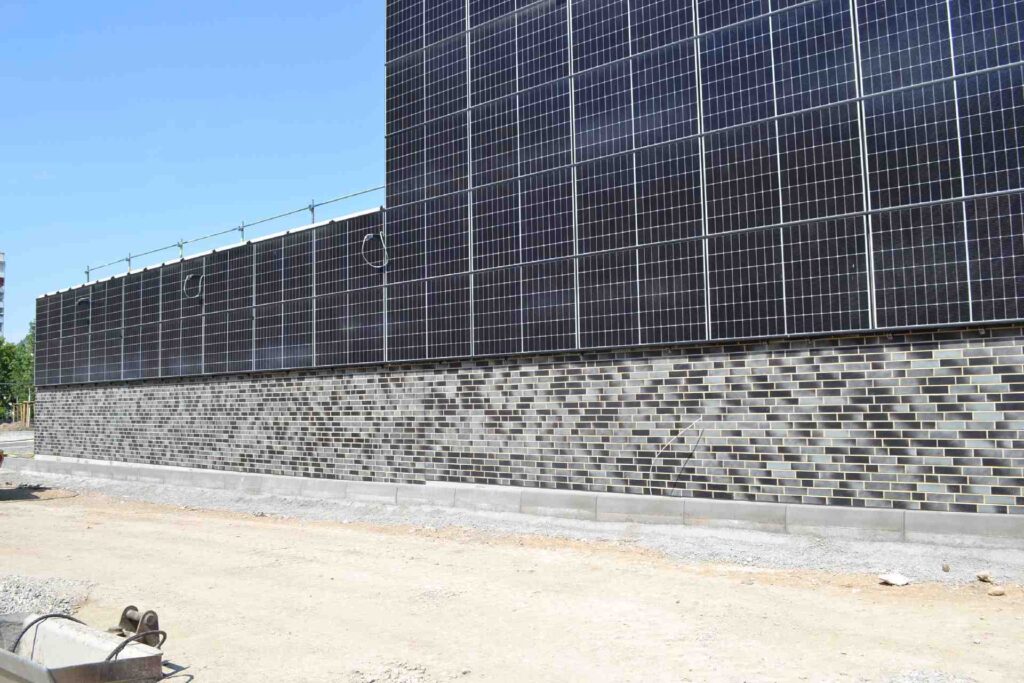 Klinkerriemchenfassade mit Solarmodulen auf einem Neubau unter blauem Himmel, Baustelle im Vordergrund.