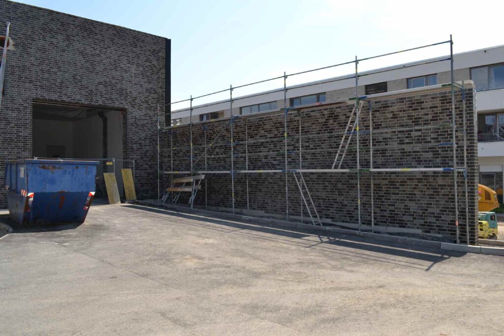 Baustelle mit Klinkerriemchenfassade und Gerüst, blauer Container und Betonfläche vor Wohngebäude