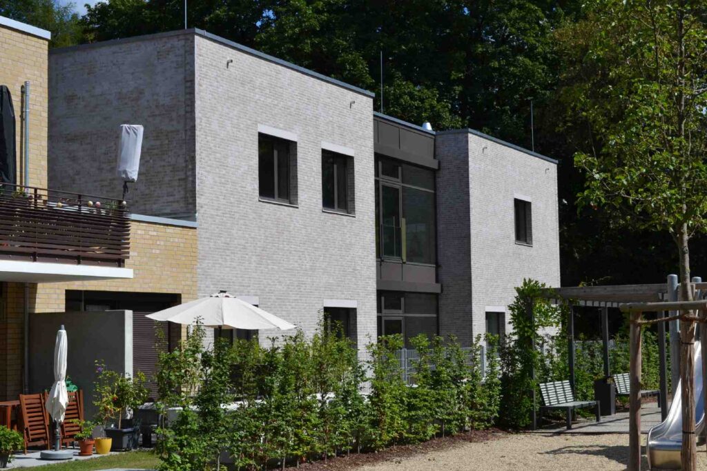 Moderne Hausfassade mit heller Klinkerriemchenfassade, großen Fenstern und gepflegtem Garten mit Terrasse.