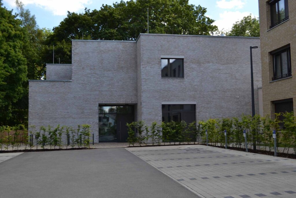 Modernes Wohnhaus mit grauer Klinkerriemchenfassade, großen Fenstern und gepflastertem Vorplatz, umgeben von Grün.