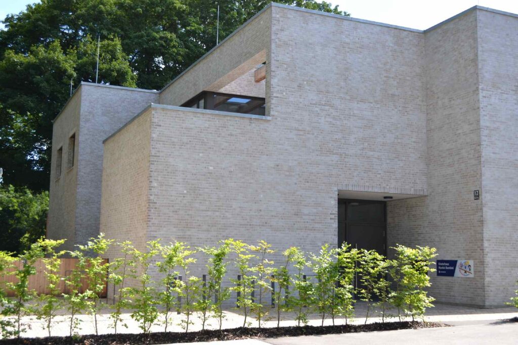 Modernes Gebäude mit heller Klinkerriemchenfassade, umgeben von jungen Bäumen und grüner Landschaft