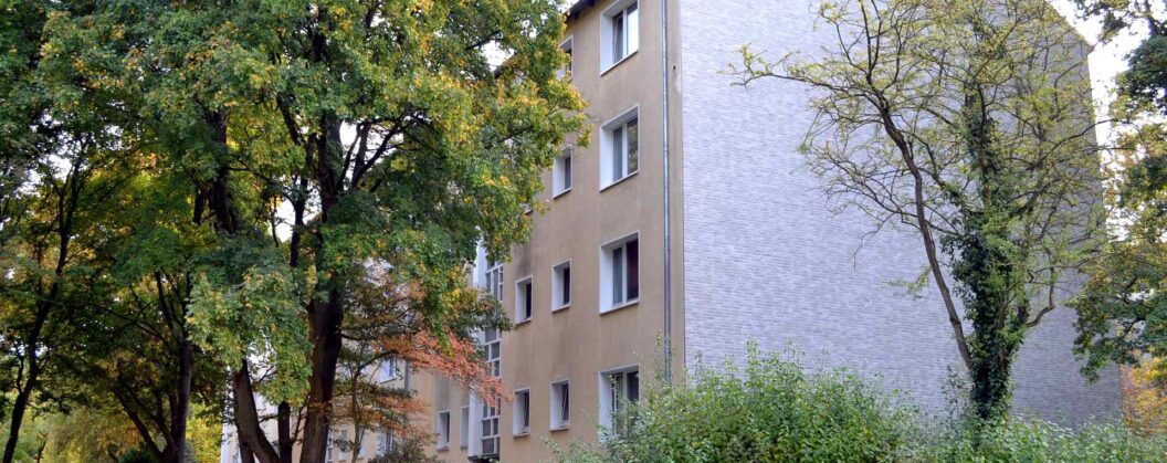 Mehrgeschossiges Wohngebäude mit beiger Hausfassade und weißer Klinkerfassade, umgeben von grünen Bäumen im Herbst.