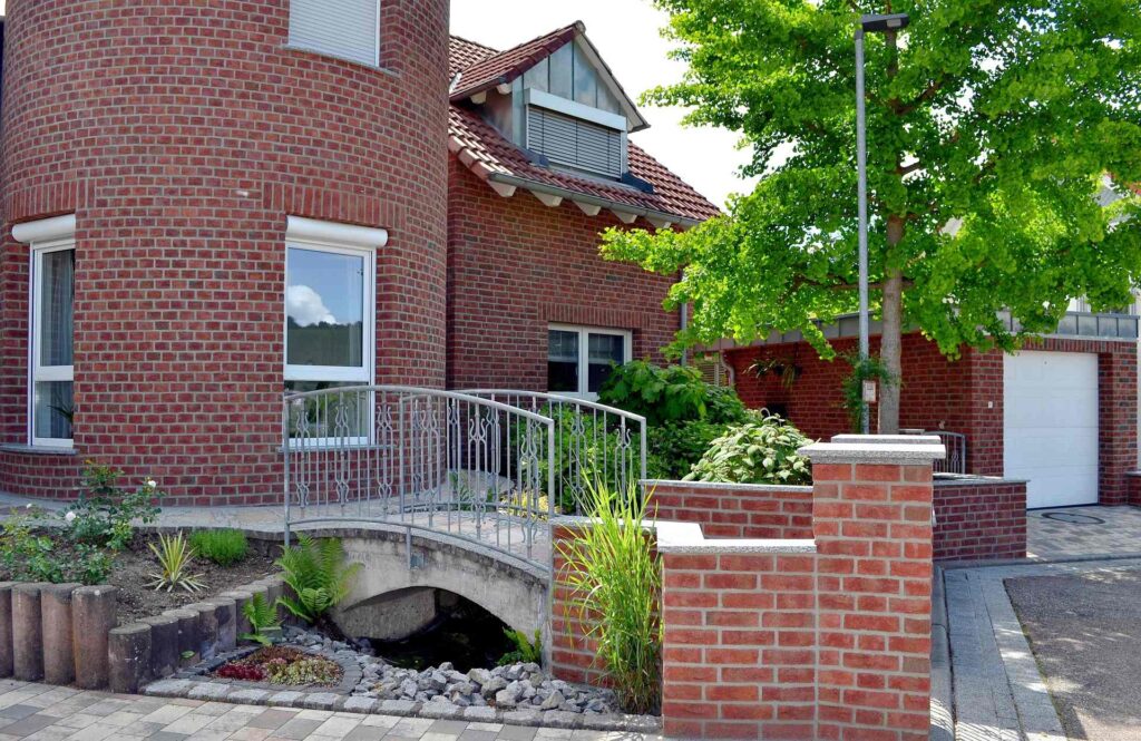 Moderní dům s klinkerovou páskovou fasádou, zahradním můstkem přes vodní prvek a zeleným záhonem