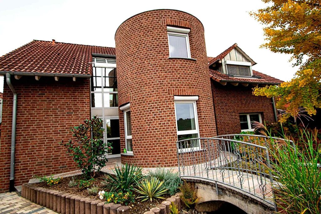 Moderní dům s kulatou věží a klinkerovou páskovou fasádou, obklopen zahradou s okrasným mostem