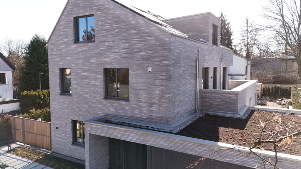 Moderní dům s fasádou z klinkerových pásků v šedém odstínu, velká okna a zelená střecha.