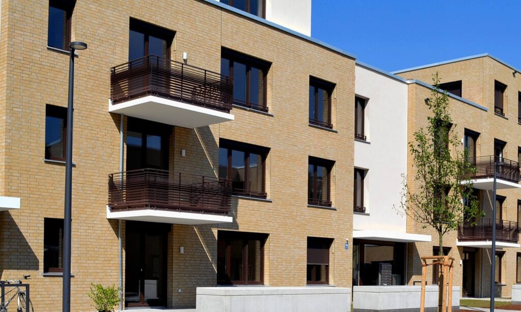 Moderní bytový dům s klinkerovou páskovou fasádou a balkony s dřevěným zábradlím pod modrým nebem.