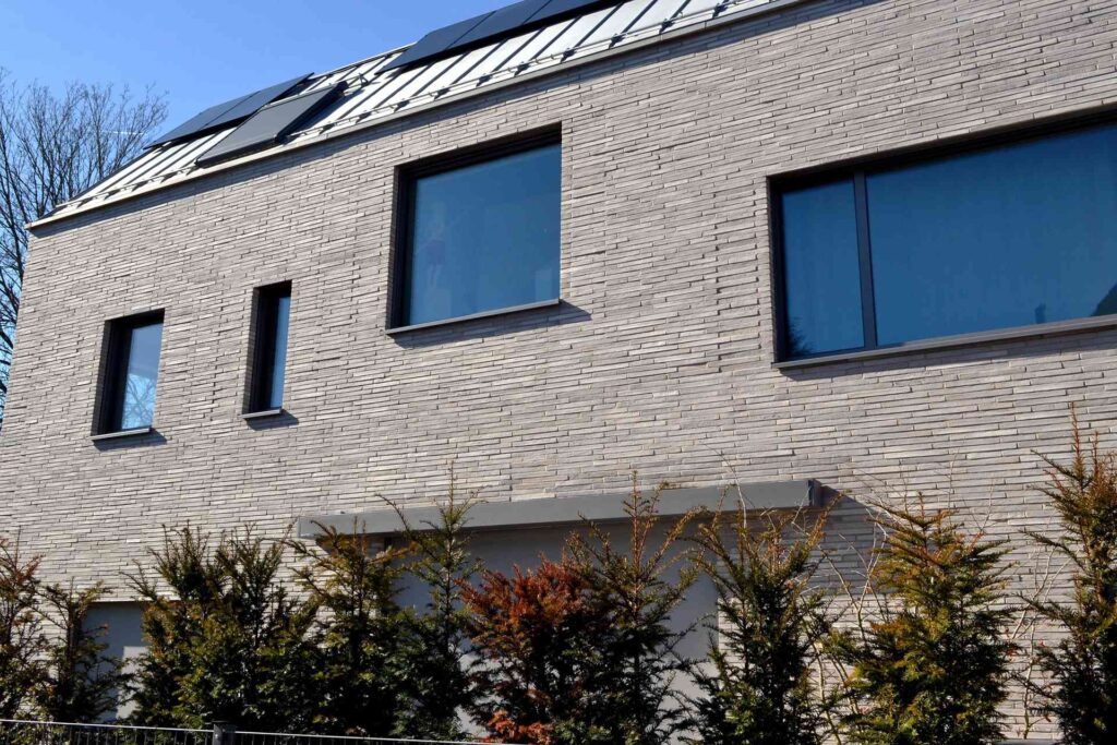 Moderní dům s klinkerovou páskovou fasádou, solárními panely na střeše a živým plotem ze smrků v popředí.