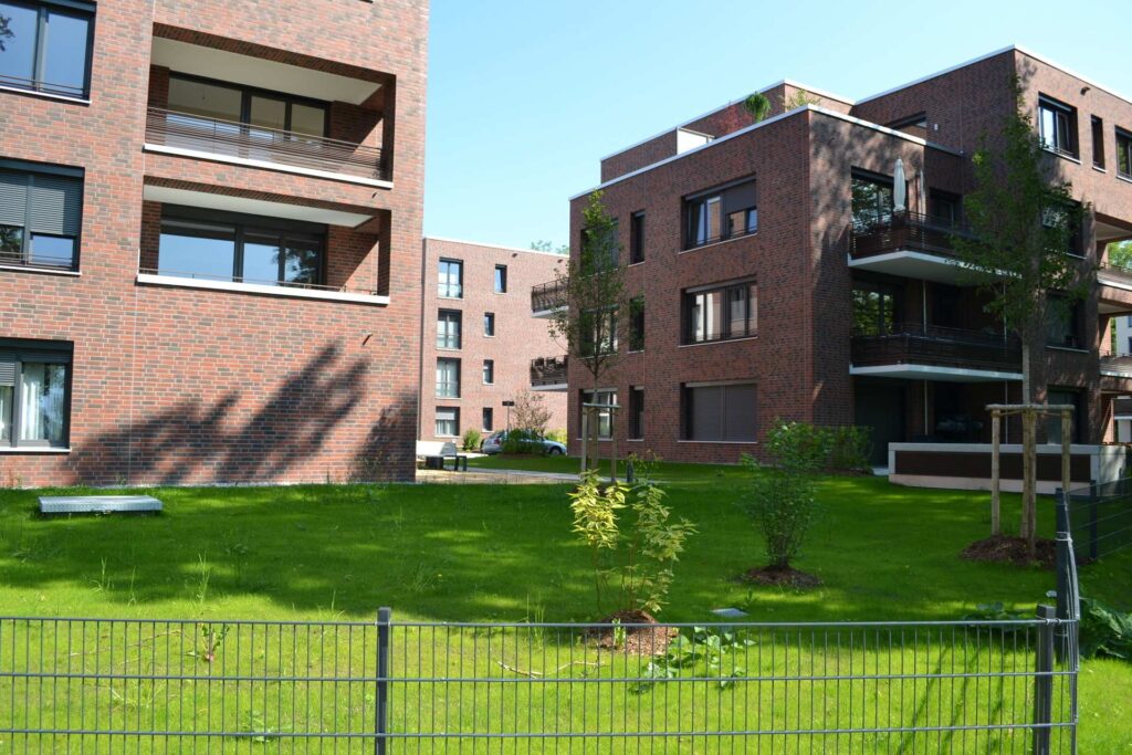 Moderní bytové domy s klinkerovou páskovou fasádou, balkony a zelenou zahradou za plotem