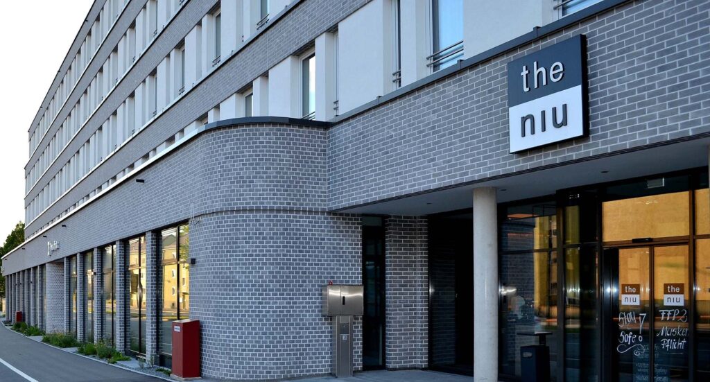 Moderní budova s klinkerovou páskovou fasádou a velkými okny, v přízemí restaurace the niu