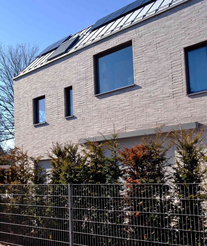 Moderní dům s klinkerovou páskovou fasádou, solárními panely na střeše a plotem s keři v popředí