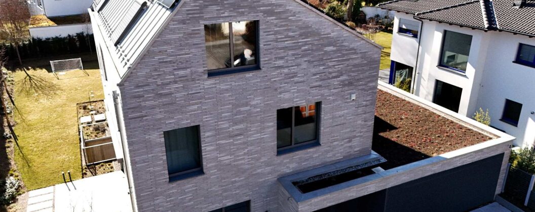 Moderní dům s tmavou cihlovou páskovou fasádou, zelená střecha a solární panely, letecký pohled