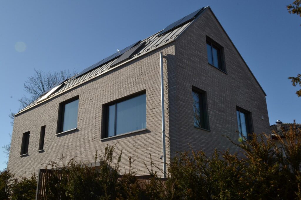 Moderní dům s klinkerovou páskovou fasádou, solárními panely na střeše a velkými okny v šedé barvě