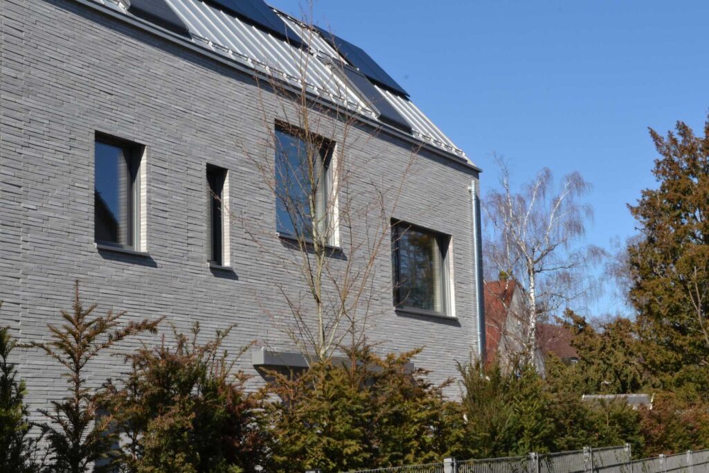 Moderní dům s fasádou z šedých klinkerových pásků a solárními panely na střeše, obklopen zelení