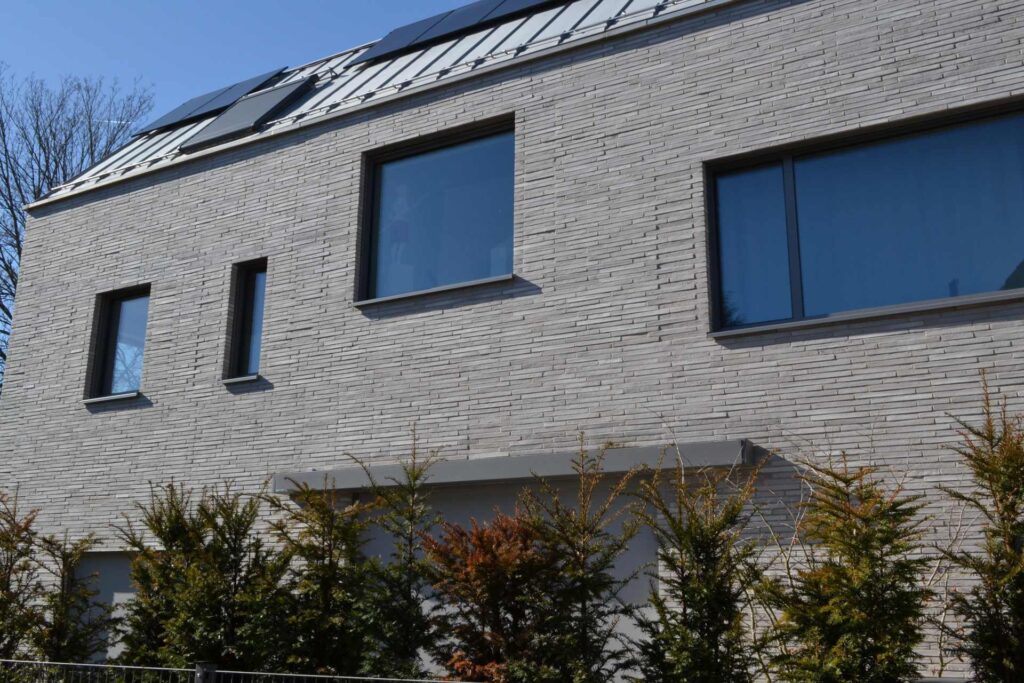 Moderní dům s fasádou z klinkerových pásků a solárními panely na střeše, obklopen zelení