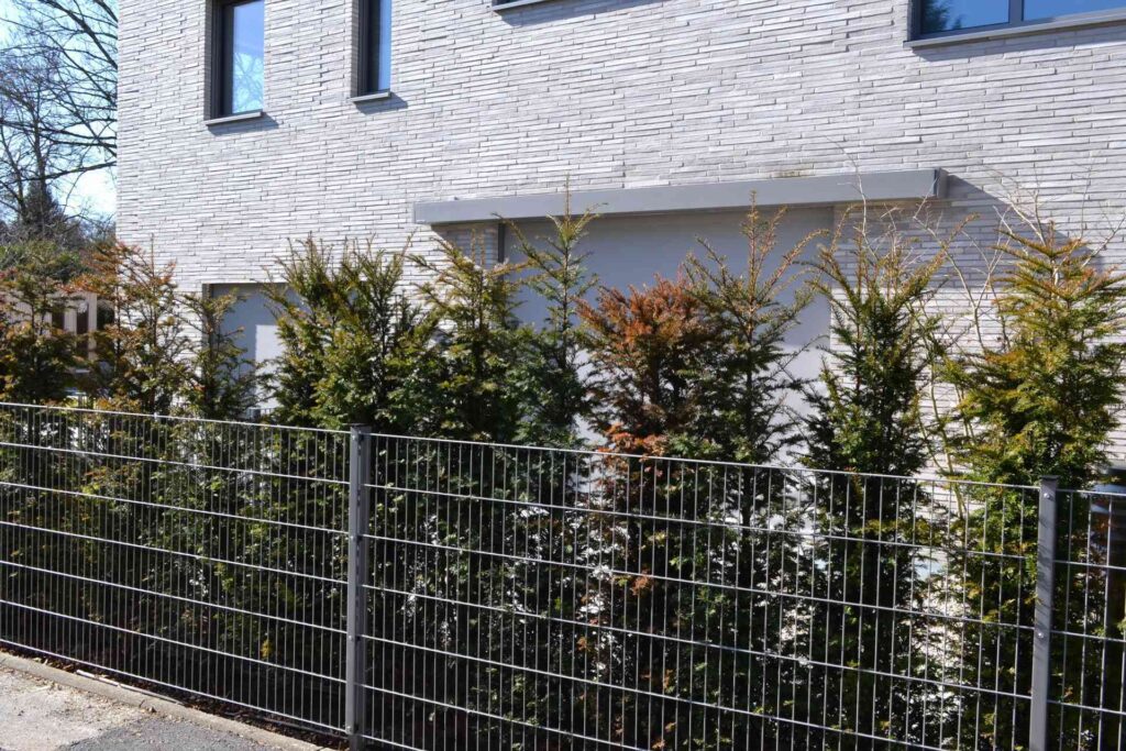 Moderní dům s klinkerovou páskovou fasádou, kovové oplocení a jehličnaté živé ploty v zahradě.