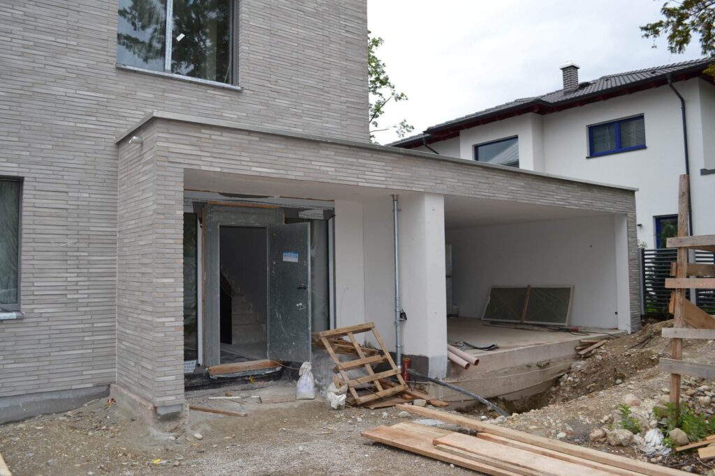 Rozestavěný dům s klinkerovou páskovou fasádou a betonovou garáží, stavební materiál rozmístěný po staveništi